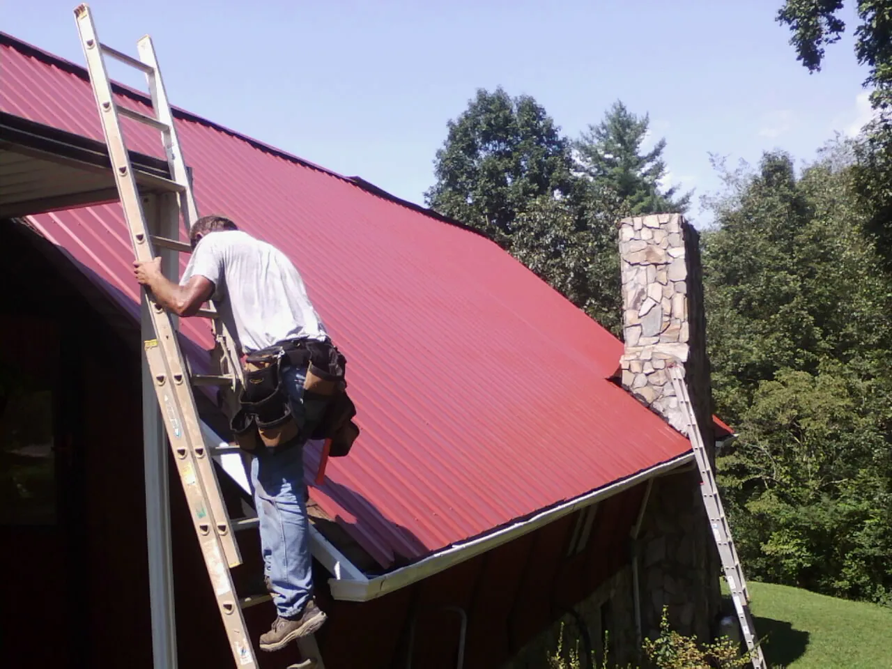 Roofer on a ladder beside a red metal roof and stone chimney — Soffit & Fascia Repair by Roofing Brothers in Neville